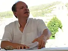 Assis à une table devant des notes, un homme en chemise blanche tourne la tête vers sa gauche.