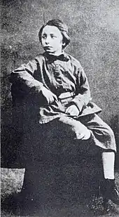 Photo en noir et blanc et de trois-quarts d’un enfant se tenant assis, le coude sur le dossier d’un fauteuil