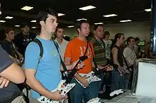 Deux personnes vues de trois-quart face, tenant des contrôleurs en forme de guitare. Ils ont le regard fixé sur un écran non-visible. Une dizaine d’autres personnes les regardent.