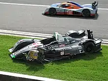 Photographie d'une voiture de sport-prototype verte, blanche, noire et rouge, vue de haut et de trois-quarts, sur de la pelouse, et d'une voiture de sport-prototype bleu et orange vue de profil