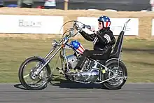 Photo d'un homme assis sur une moto au guidon démesurément grand ; son casque et le corps de la moto sont décorés aux couleurs du drapeau des États-Unis