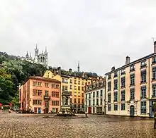 Vue d’une place avec des maisons colorées à trois étages sur deux côtés ; au fond, une colline boisée et une basilique.
