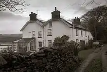 Photographie en noir et blanc de deux maisons accolées aux murs blancs, celle de gauche garnie d'une petite véranda, avec au premier plan et sur la gauche, un mur de pierres bordant le chemin d'accès ; à l'arrière plan on entrevoit un plan d'eau.