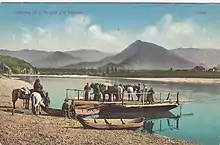 Carte postale montrant une barque à Tchouïozy sur la Katoun avant la construction du pont d'Inya. En arrière-plan se trouve un paysage montagneux verdoyant, ainsi que quelques maisons en bois sur l'autre rive. Cinq personnes et trois chevaux sont déjà sur la barque, tandis que quatre autres sont sur la berge avec leurs deux chevaux. Il y a aussi deux petites pirogues à côté de la barque