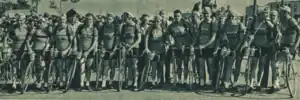 Photo en noir et blanc d'une douzaine d'hommes en tenue de cycliste, debouts à côté de leurs vélos qui se tiennent les uns à côté des autres.