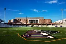 Photographie en contre-plongée d'un stade américain, vu depuis le terrain, en briques brunes sous le ciel bleu. Le symbole de l'équipe universitaire est présent au premier plan sur la pelouse.