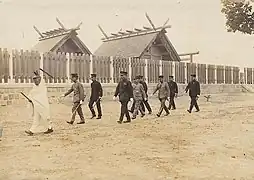 photo d'un groupe de 9 hommes en train de marcher devant le mur d'un sanctuaire. Tous portent un uniforme militaire, sauf celui en tête qui porte une tenue de prêtre shinto. Des toitures de bâtiments sont visibles derrière ce mur.