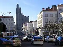 Photographie d'une église moderne en béton brut qui s'élève en haut d'une rue en pente aux immeubles bas, toits de tuiles-canal rouges, peints en blanc