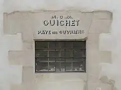 Fenêtre étroite percée dans un mur, sur lequel est inscrit «&nbsp;M.O.M. Guichet Paye des ouvriers&nbsp;».