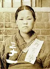 Photographie d'une très jeune femme faisant face au photographe. Elle porte un vêtement de toile grossier, et sur la clavicule d'un morceau de tissu est accroché, avec son nom écrit en kanji.