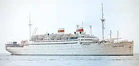 Photographie en couleurs d'un gros navire blanc avec deux cheminées. Celles-ci sont ceinturés par une ligne rouge et le logo de l'URSS. Il est vu sur une mer calme.