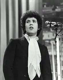 Photographie en noir et blanc avec au centre un jeune homme début trois-quart, devant un micro, costume, chemise blanche col ouvert avec un long foulard tombant, regard dirigé vers le haut à gauche, coiffure noire volumineuse frisée ; à l'arrière, à gauche grand rideau vertical, avec à droite un paysage et divers édifices