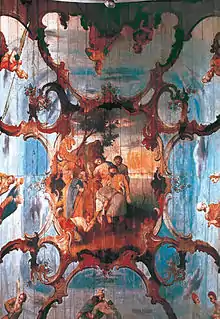 Photographie en couleurs. Gros plan sur le plafond : sur un fond de ciel bleu et blanc, de fausses moulures en trompe-l'œil soutiennent la peinture d'une scène représentant la descente de croix du Christ.