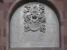 photographie en couleur montrant une niche creusée dans un mur en grès rose dont le fond en pierre grise est décoré par un relief avec des armoirie, en dessous desquelles se trouve une inscription