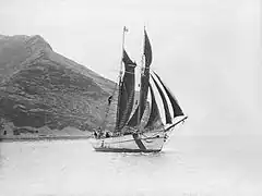 Photographie d'une goélette à deux mats dans un estuaire. En fond une île volcanique à forte pente.
