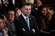 Photographie de Seth Magaziner, en costume et bras croisés, au sein de la Chambre des représentants.