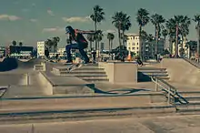 Photo d'un skater en train d'effectuer une figure de skate à Venice Beach. Il est pris en l'air durant un saut à 1 à 2&nbsp;mètres de hauteur et son skate effectue des rotations en dessous de lui. Le décor est principalement composé d'un multitude d'escaliers en béton de quelques marches. En arrière-plan assez loin, il y a des skaters et beaucoup plus loin, on aperçoit des cocotiers, la plages et des immeubles.