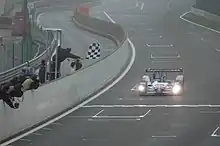 Photographie d'une voiture bleue, blanche et rouge, de face, sur une piste, devant un drapeau à damier