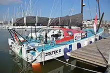 Voilier blanc amarré au ponton, vu de trois quarts arrière. Pont bleu, rouf rouge.