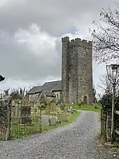 Photo d'un bâtiment allongé dominé par une grande tour carrée à créneaux d'allure massive. Une allée de gravier conduit au bâtiment derrière des rangées de pierres tombales.