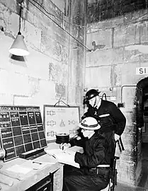 Vers la même époque, dans une salle de contrôle (où l'on distingue, contre le mur, un plan détaillé de la cathédrale) sont coordonnés des guetteurs prêts à parer aux éventuels départs d'incendies pouvant toucher le monument pendant les attaques aériennes.