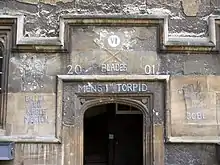 Une arche de pierre, sans porte ; au-dessus, les chiffres romains "VI" dans un cercle blanc et, à la craie, deux avirons avec des pales vertes, les mots "Blades", "2001", "Men's 1st Torpid" ; à gauche, l'inscription "Balliol SEH Keble Merton", à droite, un dragon debout tenant un drapeau au-dessus des initiales «JCBC"