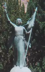 Statue de Jeanne d'ArcStatue en ange protecteur de la France, vêtue d'une robe et pourvue de deux ailes, drapeau à hampe piquée à la main, et épée au fourreau, avec un bas-relief sur le socle.