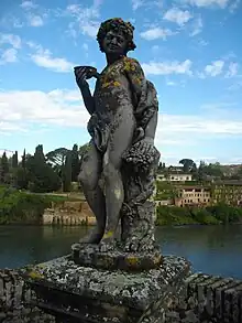 Photo couleur d'une statue de pierre marquée par le temps : lichens et mousses. Dionysos tient une coupelle de dégustation et se drape dans un tissu qui cache sa virilité. En arrière-plan, un parapet, la rivière Tarn et les jardins suspendus de la rive droite.