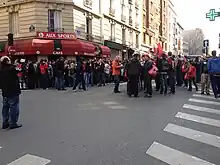 Photo montrant des personnes rassemblées dans la rue devant une enseigne rouge et noire.