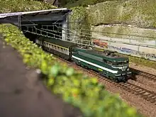 photo couleur d'une locomotive miniature sortant d'un tunnel dont les abords sont taggés. La motrice est vert foncé à bande blanche et tracte des voitures vert à bande gris métallisé au niveau des baies.