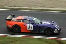 Photographie d'une voiture de grand tourisme violette, orange, noire et blanche, vue de haut et de profil, sur une piste.