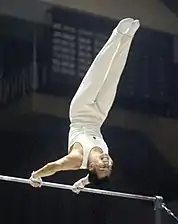 Un homme en tenue blanche, deux pieds en l'air, se tenant sur une barre.