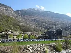 Photographie du parking de la télécabine d'Orelle avec des maisons, des bus, des automobiles garées et la télécabine : les montagnes dominent le tout.