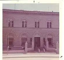 Bâtiment blanc avec des lignes horizontales sombres. Trois fenêtres et une porte au rez-de-chaussée, quatre fenêtres à l'étage.