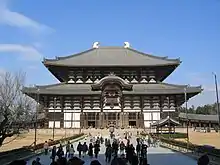 Photo couleur d'un imposant édifice religieux bouddhique en bois, à deux étages. Arrangement de poutres et de panneaux blancs, avec deux toitures de tuiles grises, sur fond de ciel bleu. Au premier plan, quelques dizaines de personnes se tiennent debout, dans l'allée qui mène aux marches de l'entrée du bâtiment.