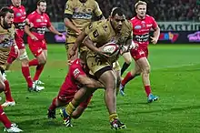 Joueur en rouge enserrant la cuisse gauche du porteur du ballon en maillot or. Derrière eux, des joueurs des deux équipes.