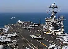 Un chasseur F/A-18 décolle du pont du porte-avions, tandis qu'un second s'apprête à décoller.