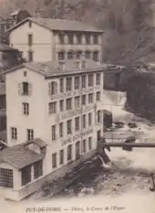 Carte postale ancienne en bistre et blanc montrant le centre de la vallée avec la Durolle juste en avant de la cascade et sur la gauche les bâtiments de l'usine d'Entraygues s'élevant en un parallélépipède de 4 étages à 7 fenêtres rectangulaires par niveau. On lit très clairement sur les ceintures de maçonnerie entre les étages de haut en bas : Manufacture de Coutellerie Fine, Sauzede Père et Fils, Usine d'Entraygues