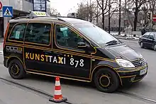 Photo d'une voiture noire avec des lignes jaunes, sur lequel est posée une plaque de taxi, traversant une ligne de tramway.