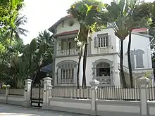 Façade d'une maison blanche à deux étages, entourée de palmiers.