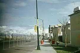 Une rue déserte bordée de lampadaires. Au fond, on devine les montagnes. À droite, des petits bâtiments de deux étages s'enchaînent.