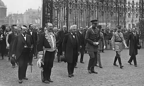 Photo en noir et blanc d’un groupe d’hommes (civils et militaires) pénétrant dans une cour entourée d'une grille