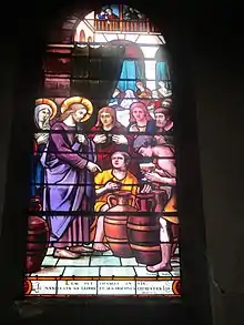 Vitrail représentant Jésus auréolé pointant son doigt vers des tonneaux, entouré d'une petite foule de personnages secondaires.