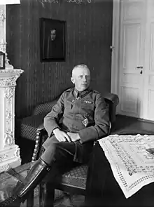 photo en noir et blanc d'un homme assis dans un salon, il porte un uniforme et des bottes en cuir, au fond on distingue un tableau sur un mur et une porte blanche