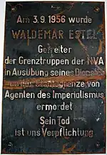Plaque de métal endommagée indiquant «&nbsp;Am 3.9.1956 wurde WALDEMAR ESTEL Getreiter der Grenztruppen der NVA in Ausübung seine Dienstes an der Staatsgrenze von Agenten des Imperialismus ermordet. Sein Tod ist uns Verpflichtung.&nbsp;»