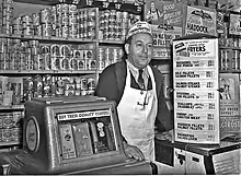 Photographie en noir et blanc d’un épicier souriant, cravaté, portant un tablier blanc à bretelle unique passant derrière le cou, coiffé d’un calot publicitaire ; l’homme s’appuie à une machine distributrice de café et à un comptoir surmonté de panneaux publicitaire ou annonçant les prix de marchandises. Derrière lui, l’étagère qui court tout le long du mur est remplie d’une multitude de boîtes de conserve, parfois cachées par des calicots publicitaires supplémentaires, donnant l’impression qu’il n’y a plus 1&nbsp;cm2 de disponible.