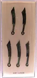 Photographe montrant cinq objets en forme de couteau derrière une vitrine. De formes similaires, les objets sont divisés en deux rangées de 2 et 3 couteaux.