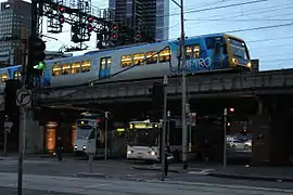 Un tramway à côté d'un bus Melbourne Bus Link, avec un métro Trains X'Trapolis 100 passant au-dessus.