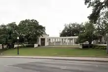 Une construction blanche sur un monticule gazonneux, entourée d'arbres. Au premier plan une route