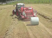 Dans cette photographie prise dans un champ, outre la balleuse et la botte, se distingue également un tracteur.